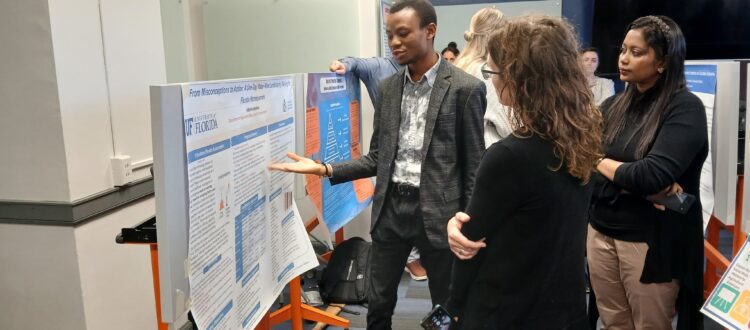 Adeyinka Ayodele with advisor Dr. Larua Warner presenting his research on a poster. There are other presenters in the background.