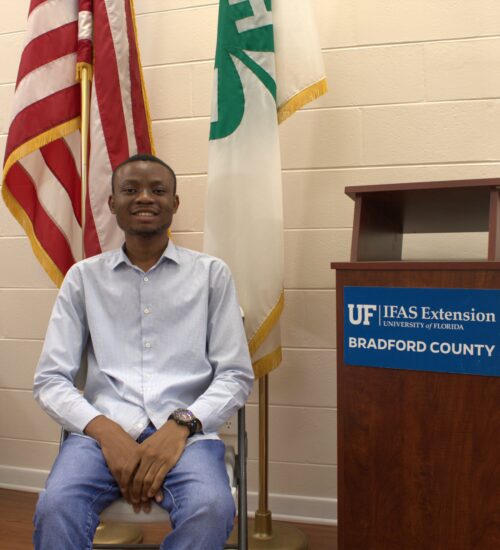 Adeyinka Ayodele sitting in front of two flags and next to a podium with the UF/IFAS Extension Bradford County logo.